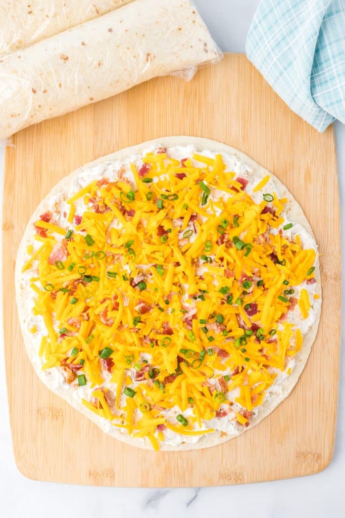 Cheddar cheese, green onion, bacon and cream cheese mixture with chicken spread on a tortilla from overhead on a cutting board.