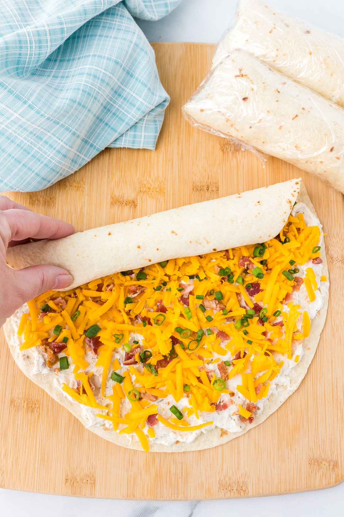 Tortilla loaded with cheddar cheese, bacon, green onions and a cream cheese mixture being rolled tightly.