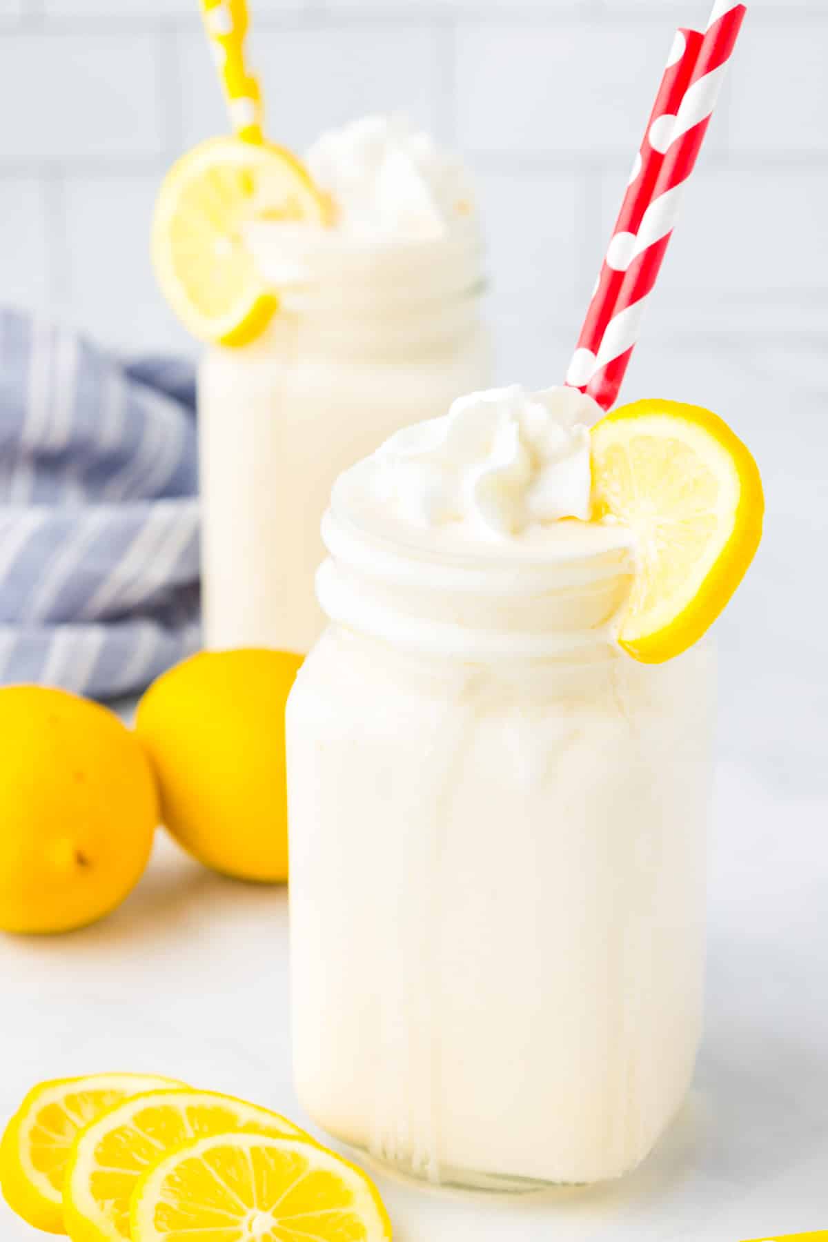 Two frosted lemonades in glass jars topped with whipped cream and a lemon slice with more lemons nearby on the counter.