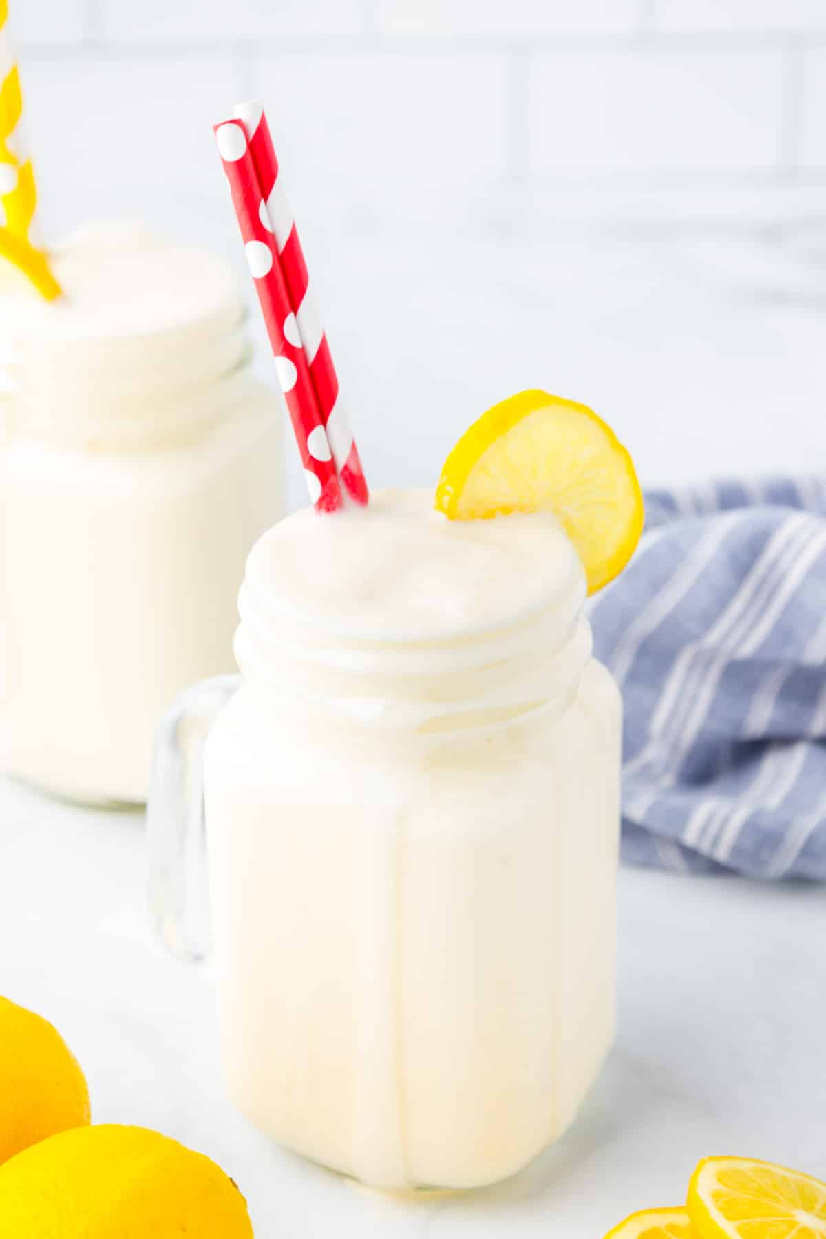 Side view of a glass jar with a handle full of frosted lemonade with a dribble down the side, topped with a fresh slice of lemon and red and white straws. . One more frosted lemonade is in the background.