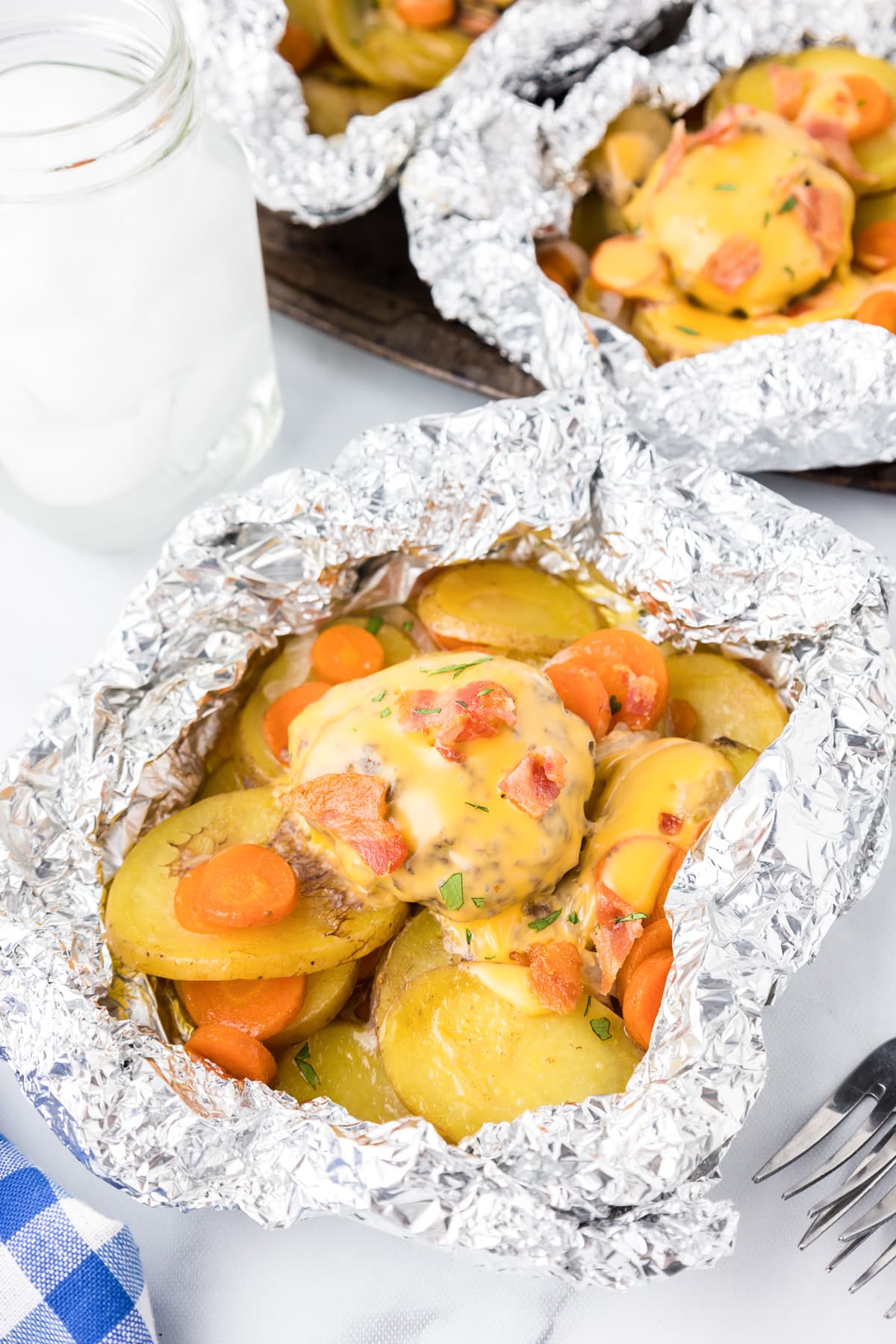 Open foil with potatoes, onions, carrots and a burger covered in cheese and chopped bacon on a table.