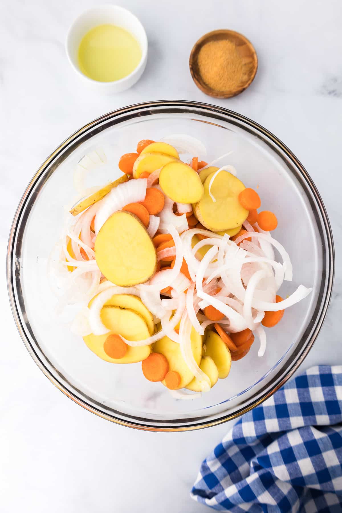 A large bowl of mixed thinly sliced potatoes, onions and carrots with oil and seasoned salt in small bowls nearby.