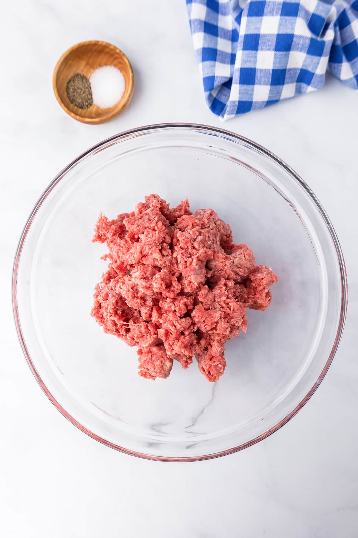 Ground beef in bowl with salt and pepper and a napkin on the counter nearby.