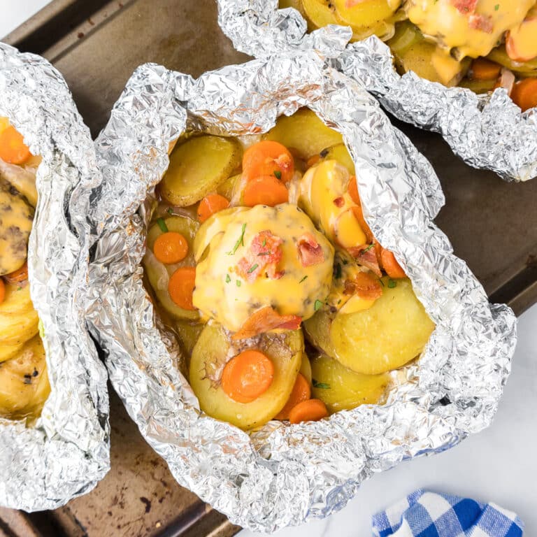 Close up a three rolled foil packets from above full of thin sliced potatoes, carrots, onions and topped with a burger with bacon.