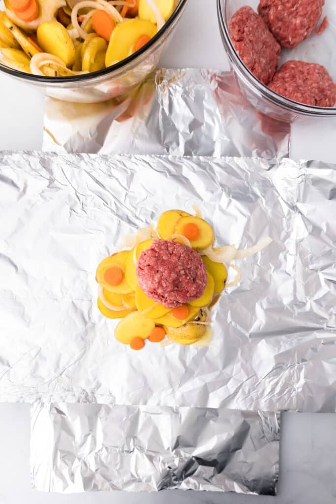 Two piece of foil laid in a large plus sign with vegetables piled high and topped with a raw burger in the middle of the foil.