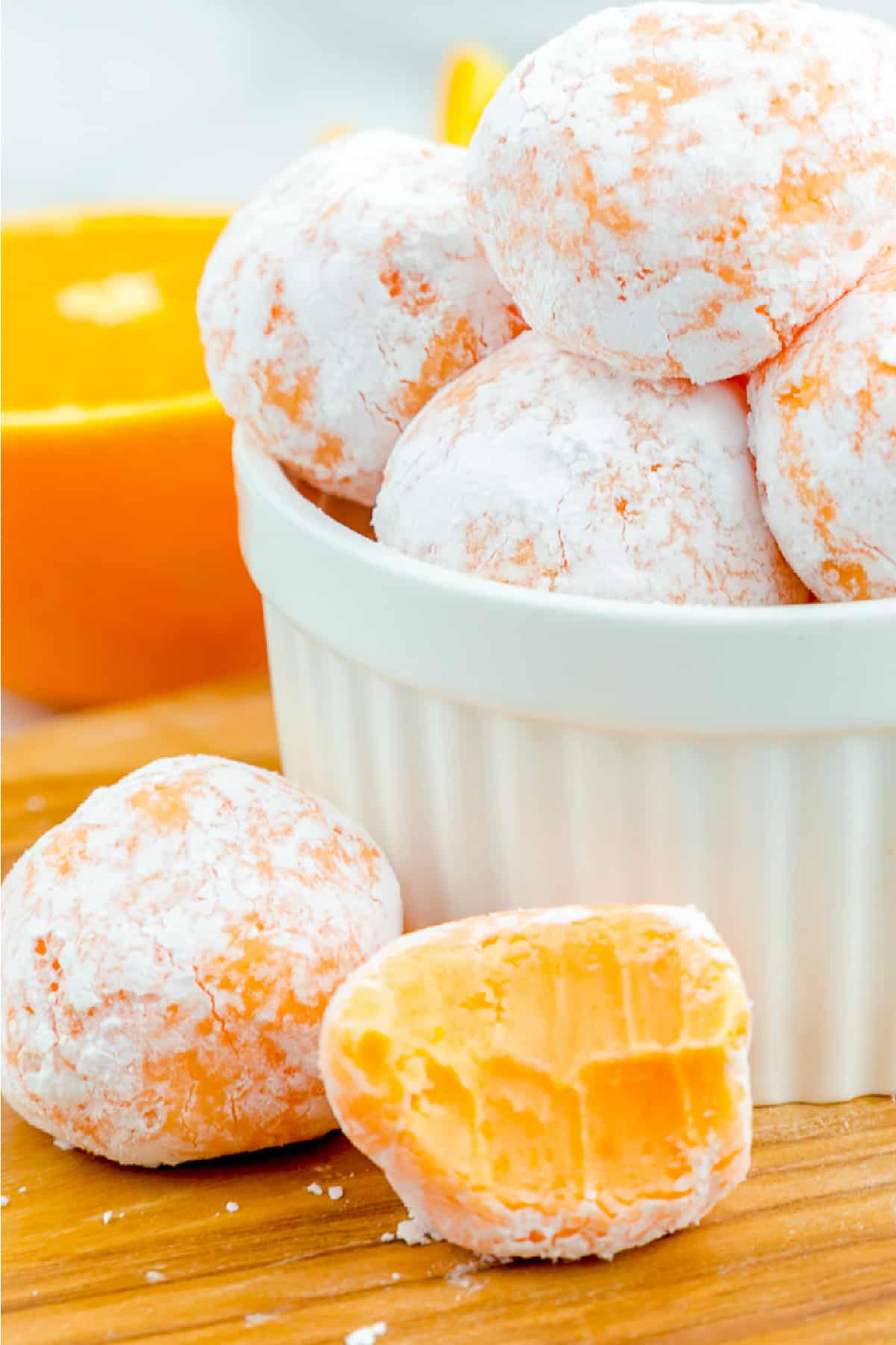 Cropped in orange creamsicle truffle missing a bite next to a bowl stacked with more truffles covered in powdered sugar.
