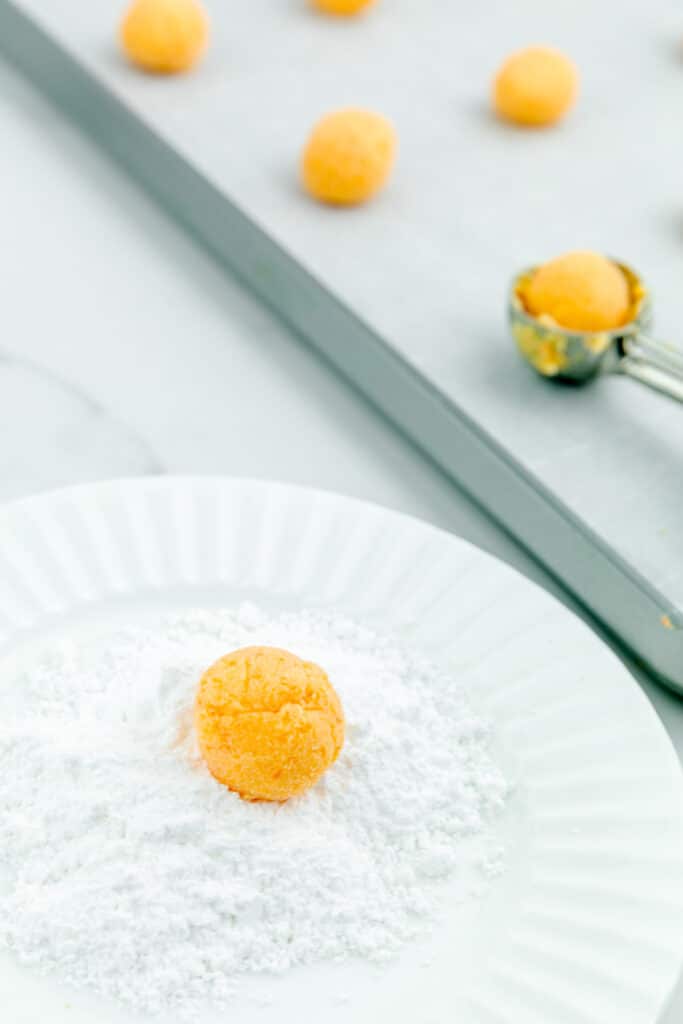 Orange creamsicle truffle being rolled on a plate of powdered sugar with a scoop and more truffles behind on the counter on a pan.