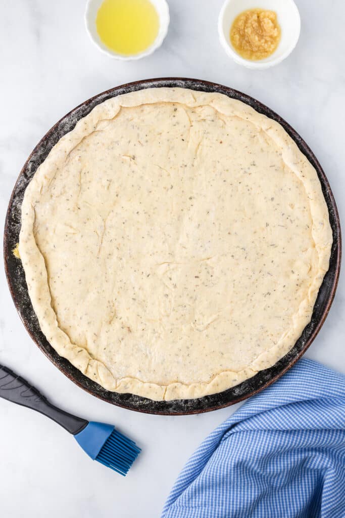 Pizza dough stretched on a pan with the crust rolled at the edges, with olive oil, garlic and a silicone brush on the counter nearby.