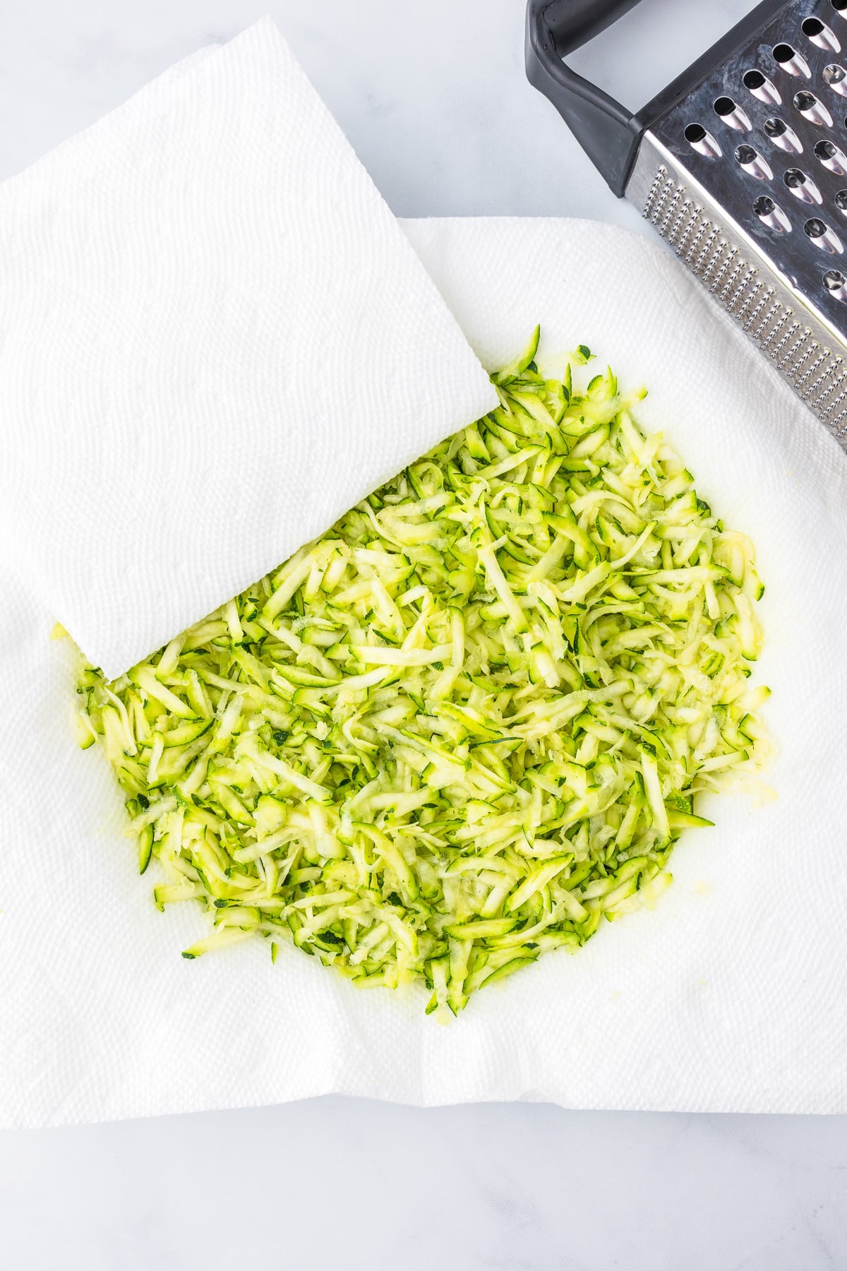 Shredded zucchini being dried on paper towels with a metal grater on the counter nearby.