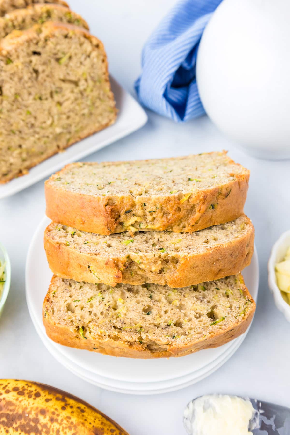 Zucchini banana bread slices on a plate piled high with more on a platter nearby on the counter and other items such as butter and a pitcher nearby.