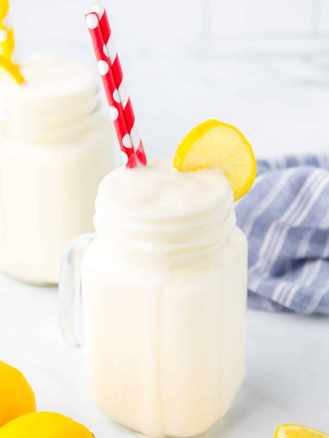 Side view of a glass jar with a handle full of frosted lemonade with a dribble down the side, topped with a fresh slice of lemon and red and white straws. One more frosted lemonade is in the background.