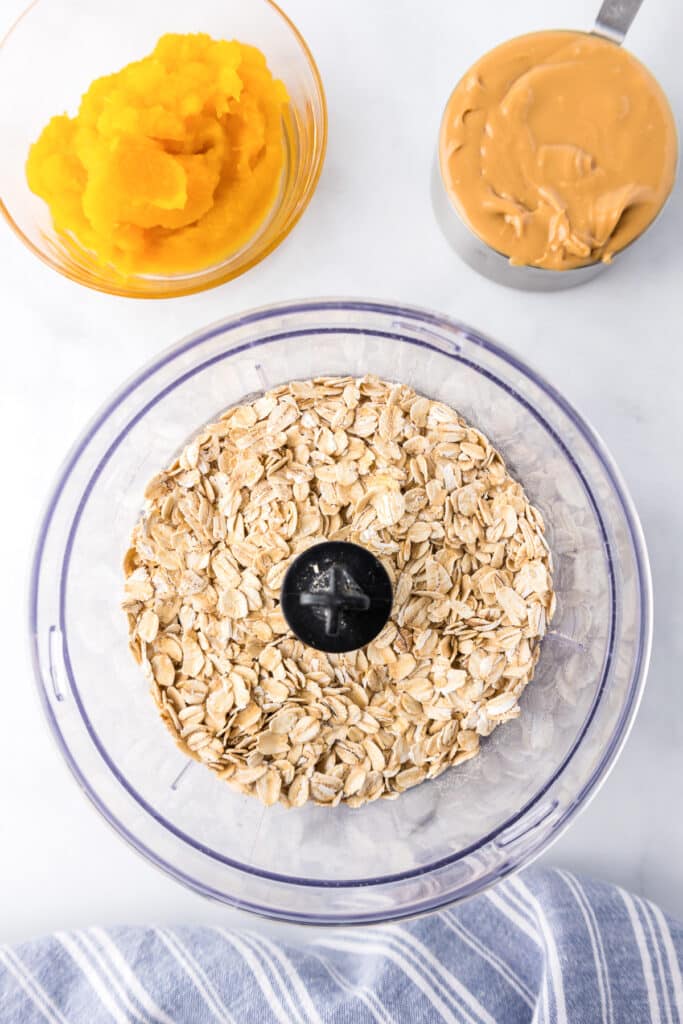 Oats in a food processor from overhead with a measuring cup of peanut butter and a bowl of pumpkin nearby.