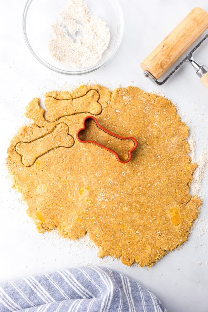 Bone shaped cookies being cut out from a peanut butter dough with a rolling pin and a bowl of pulsed oats nearby.