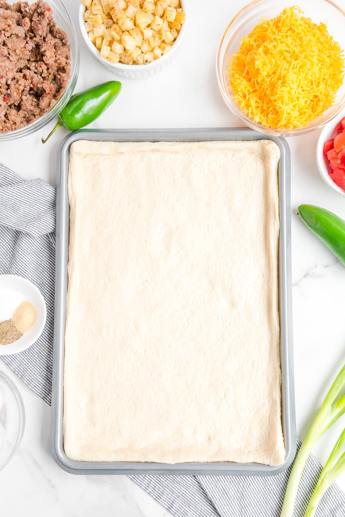 Pizza dough stretched over a baking sheet to make a rectangular pizza from above with bowls of other pizza ingredients nearby.