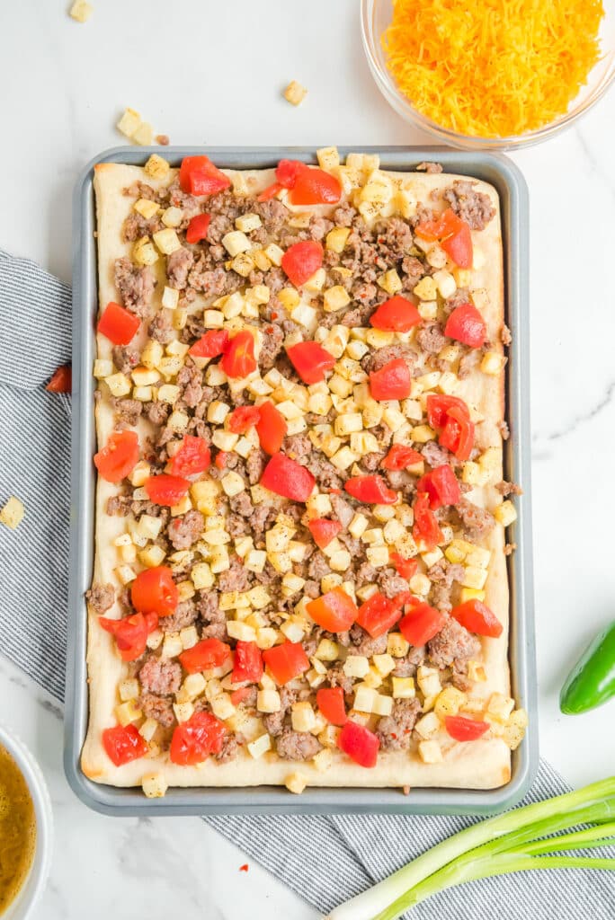 Pizza crust in a baking sheet from above topped with sausage, diced potatoes and tomato pieces from above.