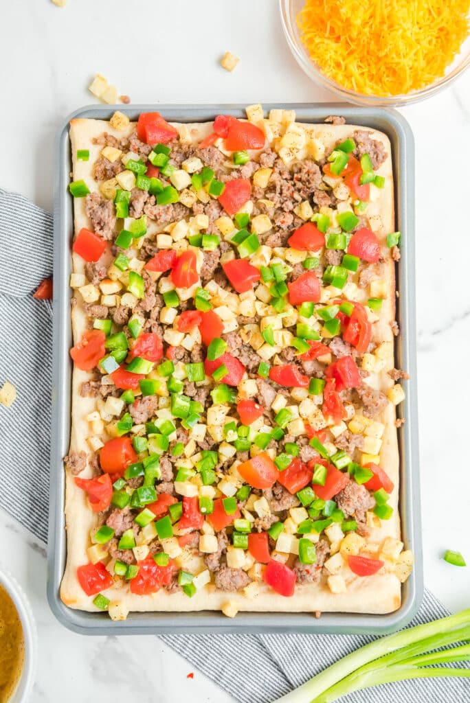 Green onions, tomatoes, diced potatoes and sausage sprinkled across a pizza crust on a baking sheet from above.
