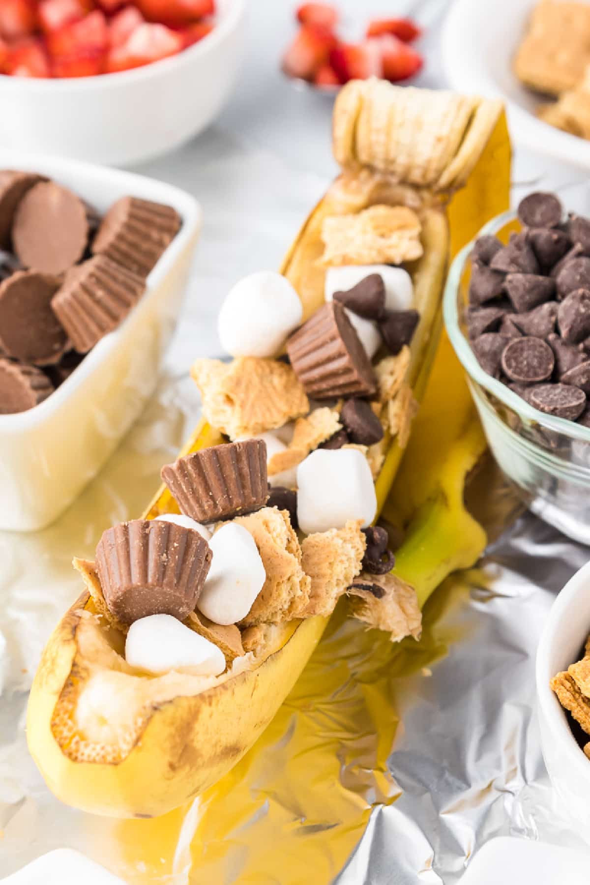 Single banana boat stuffed with chocolate chips, mini marshmallows, peanut butter cups and graham cracker pieces with more toppings in bowls nearby.