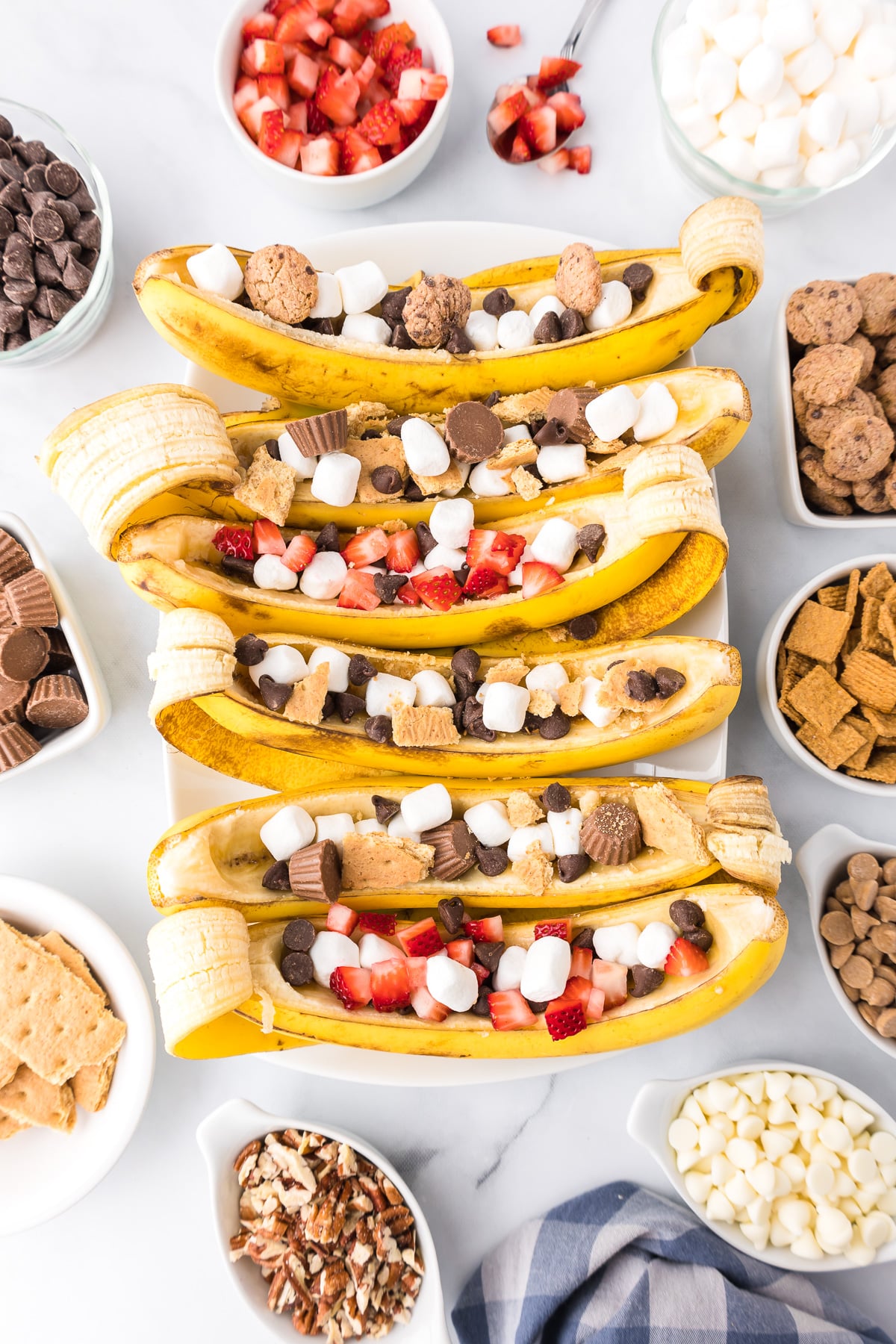 Six bananas hollowed out in a row filled with toppings like chocolate, marshmallows, peanut butter cups and strawberries pieces with more toppings in bowls nearby.