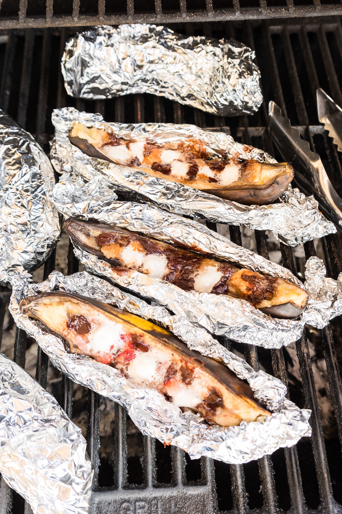 Bananas wrapped in foil and stuffed with chocolate, marshmallows and strawberries on a grill with some of them unwrapped to see the gooey inside.