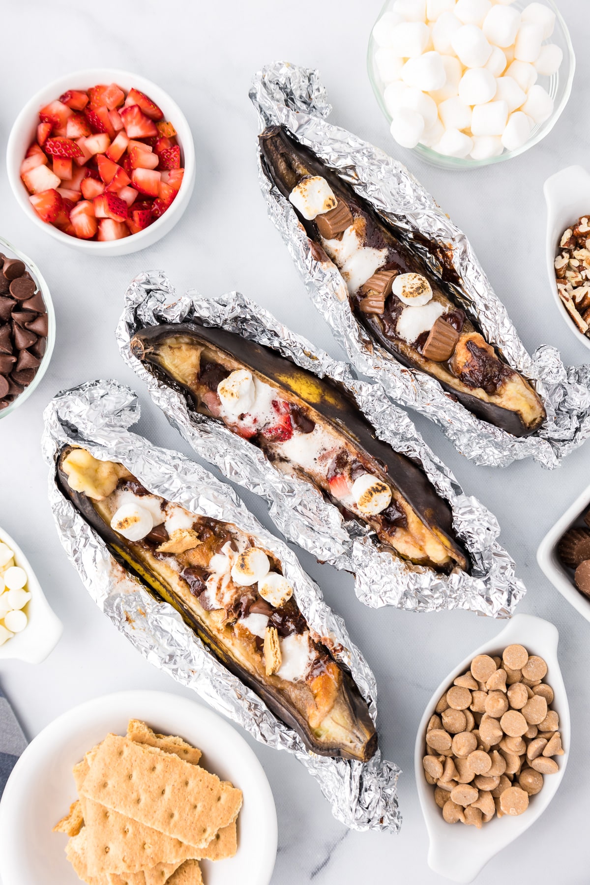 Three cooked bananas stuffed with sweet toppings with aluminum foil peeled back and more toppings around the bananas.
