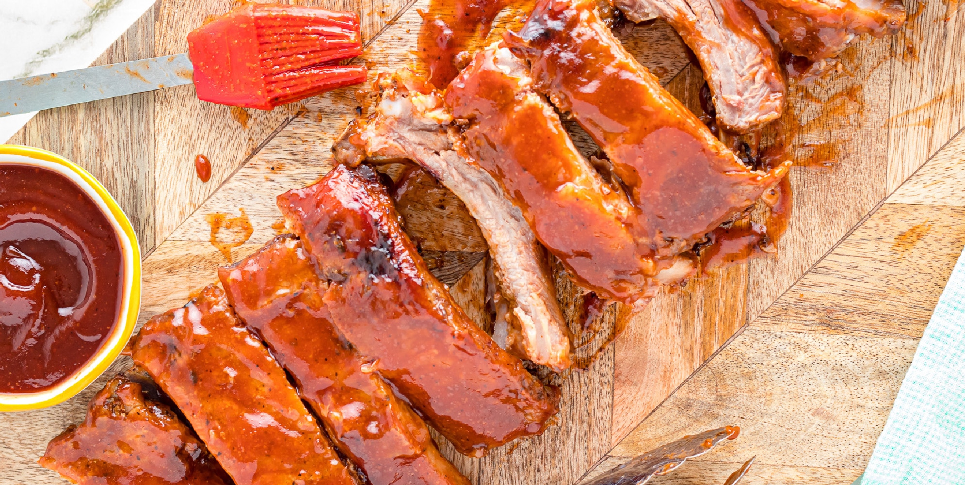 Sliced ribs on a cutting board slathered in sauce next to a brush from overhead.