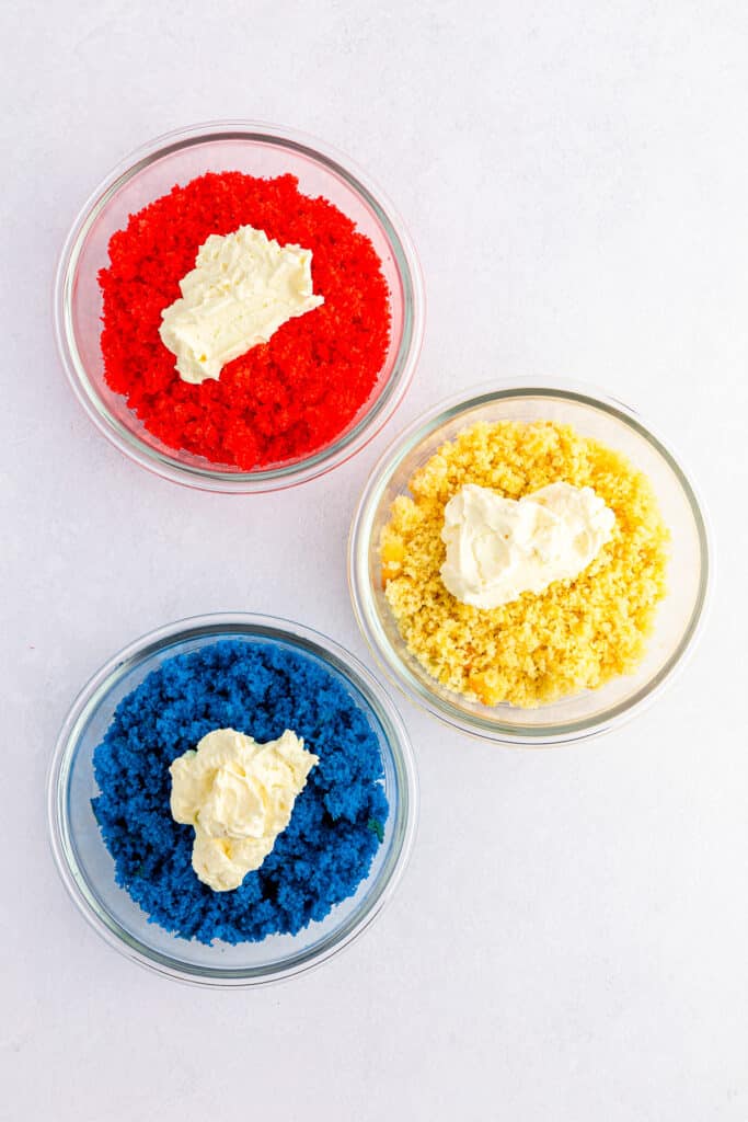 Cream cheese mixture on top of each of the three bowls of cake crumbs which are dyed red, white and blue.