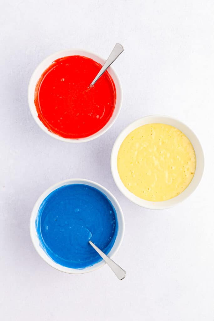 Cake batter in three bowls being mixed with food coloring with spoons to make one red, one blue and one white bowl of batter.