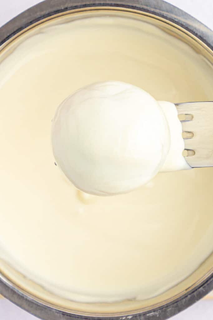 Cake ball being dipped in white chocolate in a bowl from above close up.