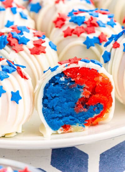Close up of the inside of a red and blue cake ball next to more cake balls on a plate covered in red and blue star sprinkles and white chocolate.