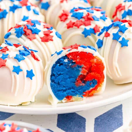 Close up of the inside of a red and blue cake ball next to more cake balls on a plate covered in red and blue star sprinkles and white chocolate.