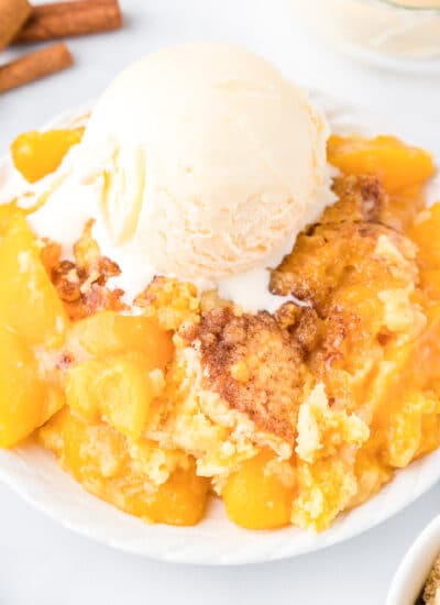 Peach dump cake gooey on a plate topped with a scoop of vanilla ice cream from the side on a counter.
