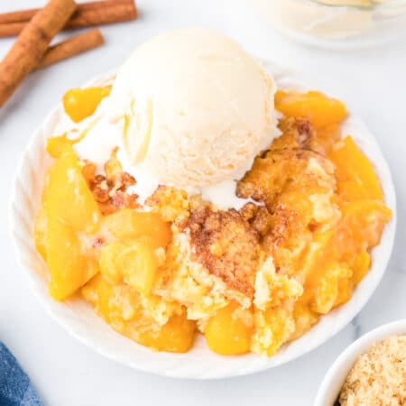 Peach dump cake gooey on a plate topped with a scoop of vanilla ice cream from the side on a counter.