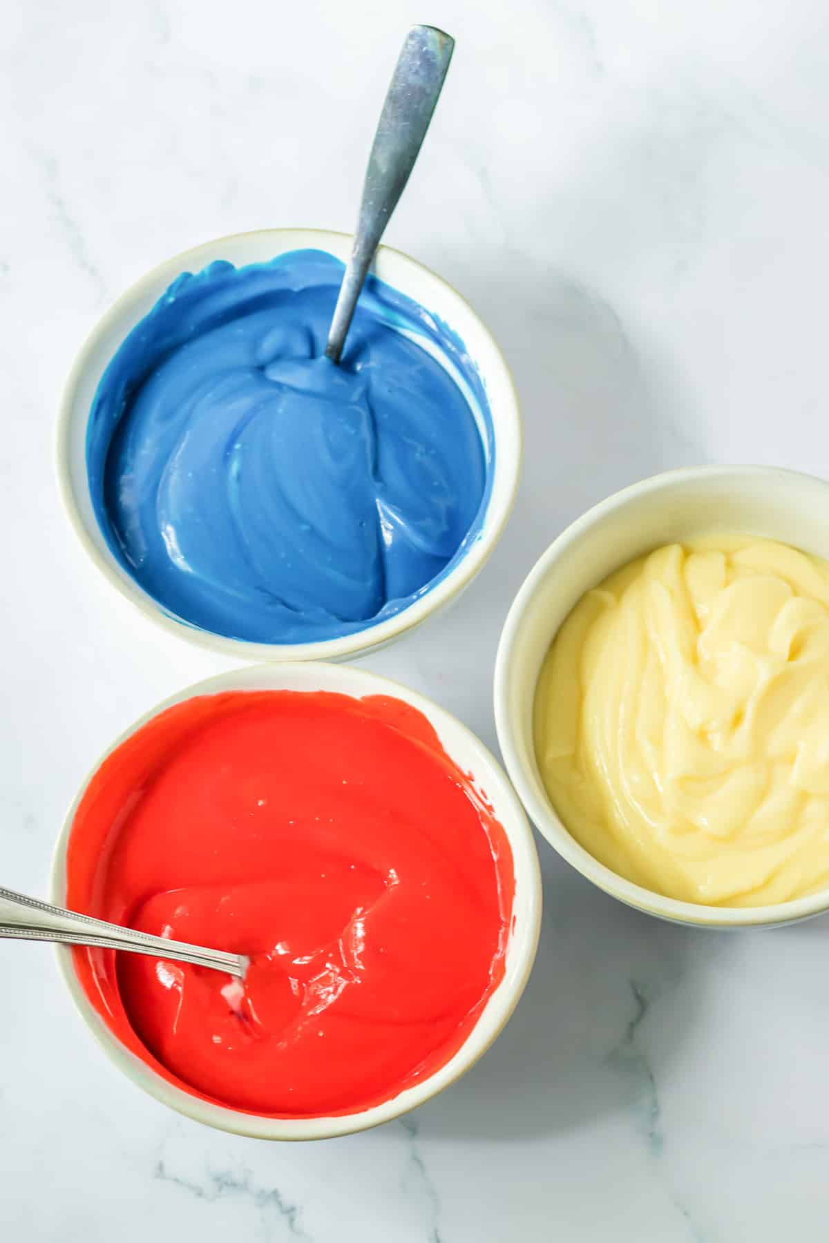 Blue, red and white pudding in three different bowls being mixed with spoons.