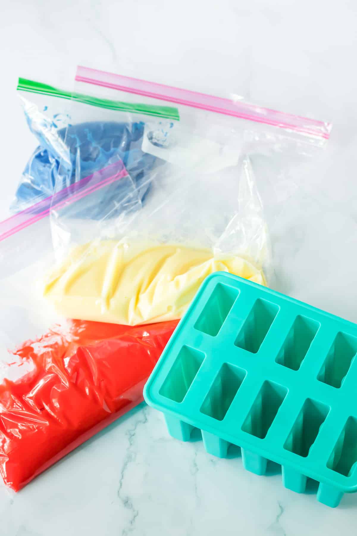Blue, white and red pudding each in a zip top bag next to a silicone popsicle mold on a kitchen counter.