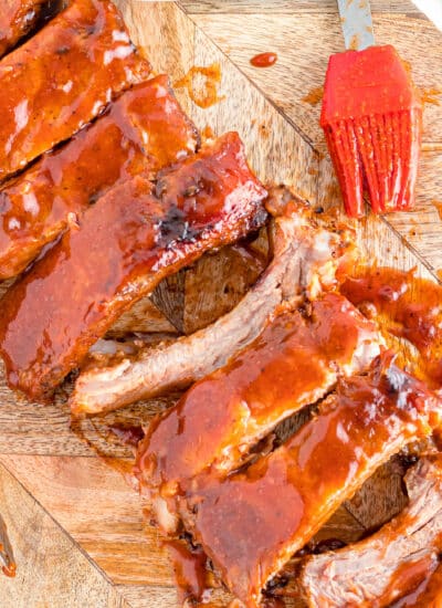 Sliced saucy Dr. Pepper Ribs on a cutting board at an angle with a silicone brush full of sauce next to the ribs from overhead.
