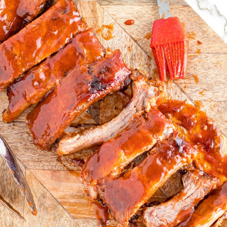 Sliced saucy Dr. Pepper Ribs on a cutting board at an angle with a silicone brush full of sauce next to the ribs from overhead.