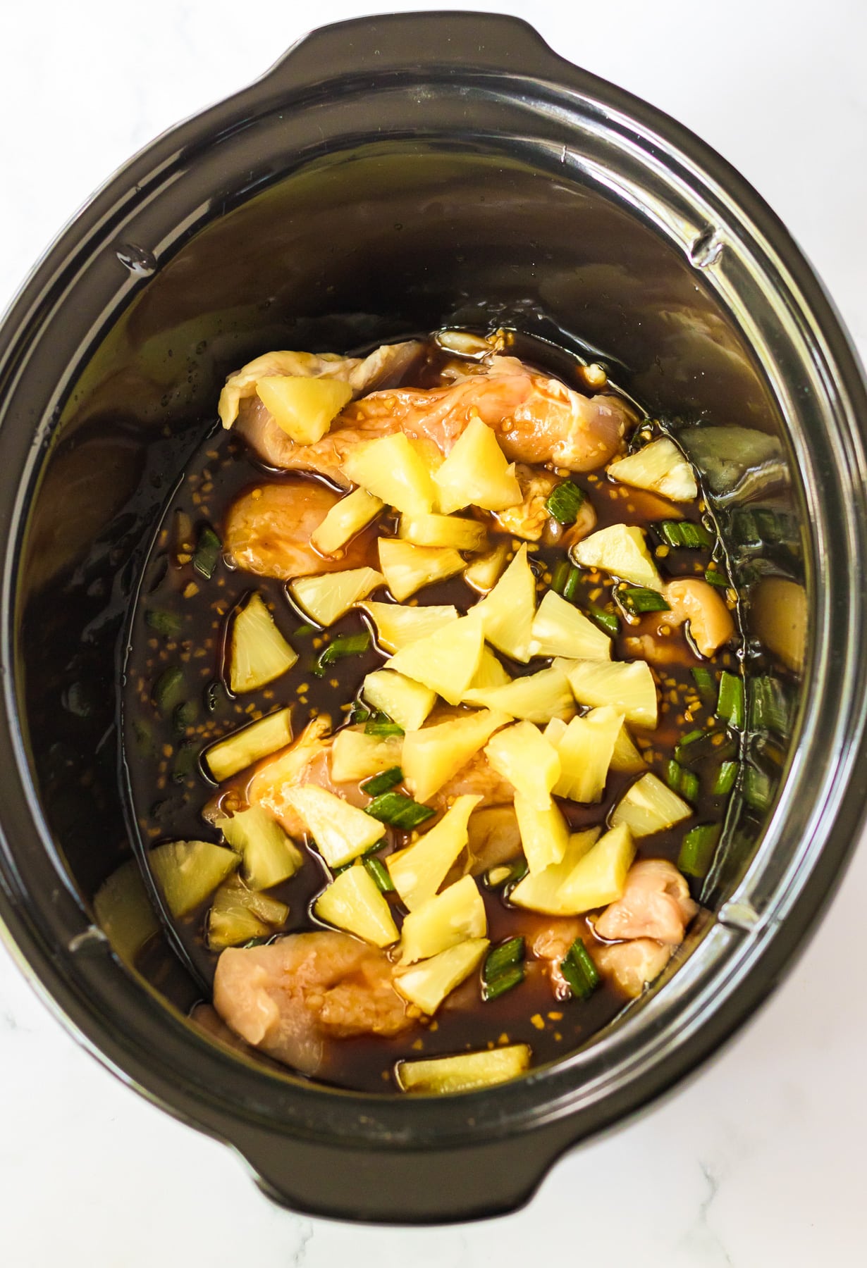 Raw chicken and pineapple pieces in a sauce in a slowcooker base from overhead.