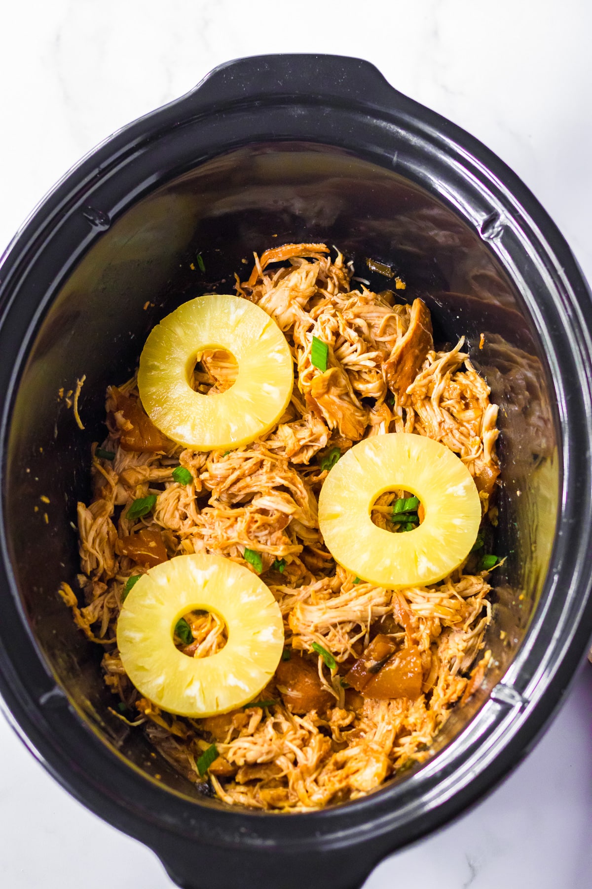 Shredded chicken in the slow cooker base with three rings of pineapple slices on top.