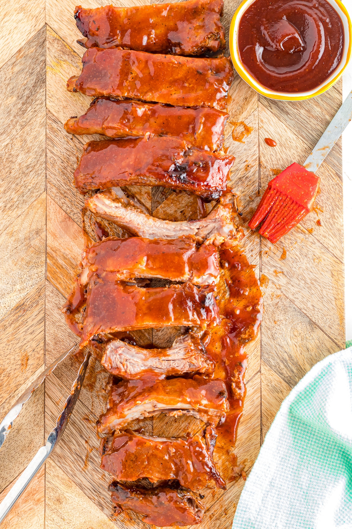 Close up of ribs sliced and covered with BBQ sauce on a cutting board with a bowl of sauce and a brush nearby.