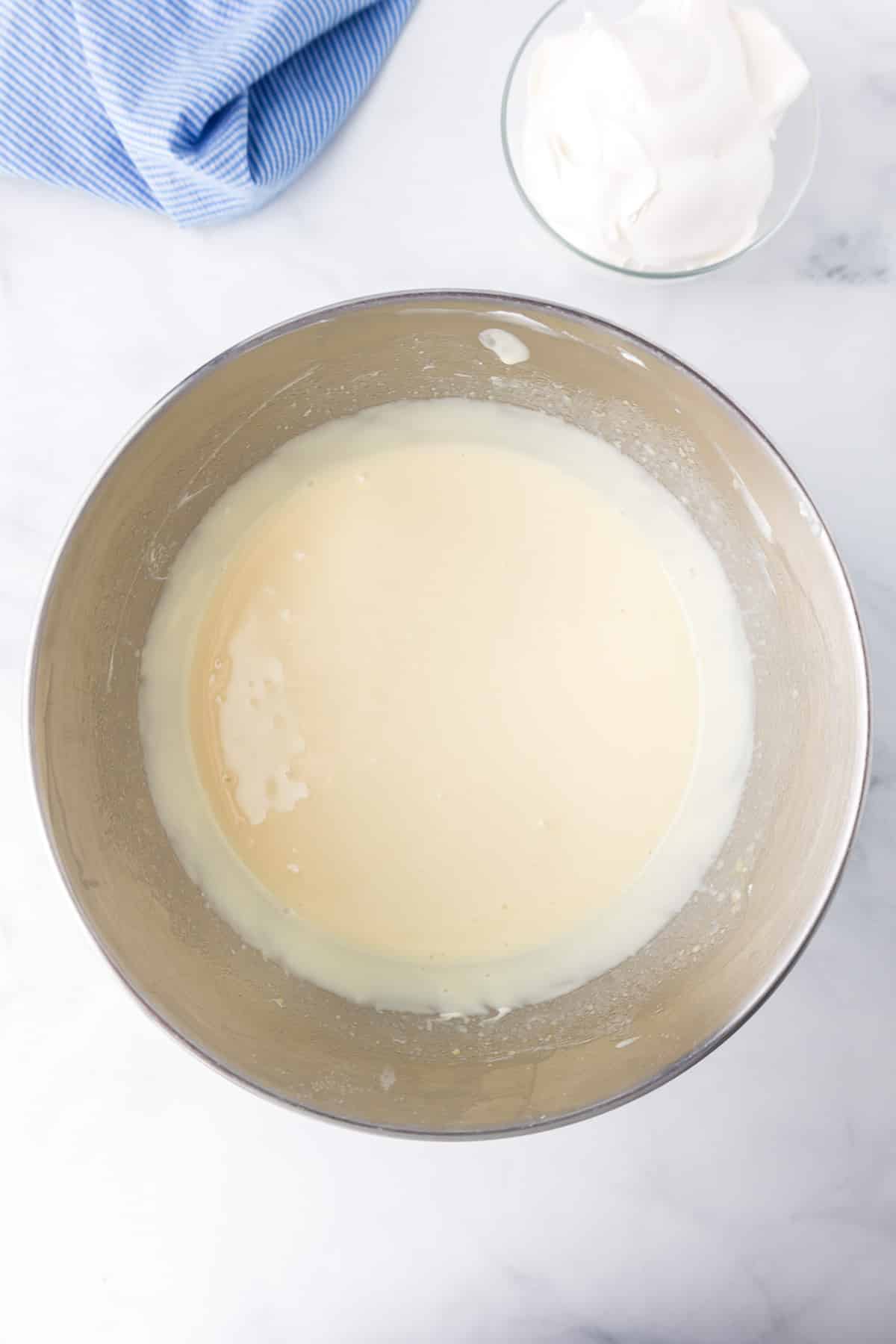 Pudding mixture and milk being blended into the mixing bowl with whipped topping in a bowl on the counter nearby.