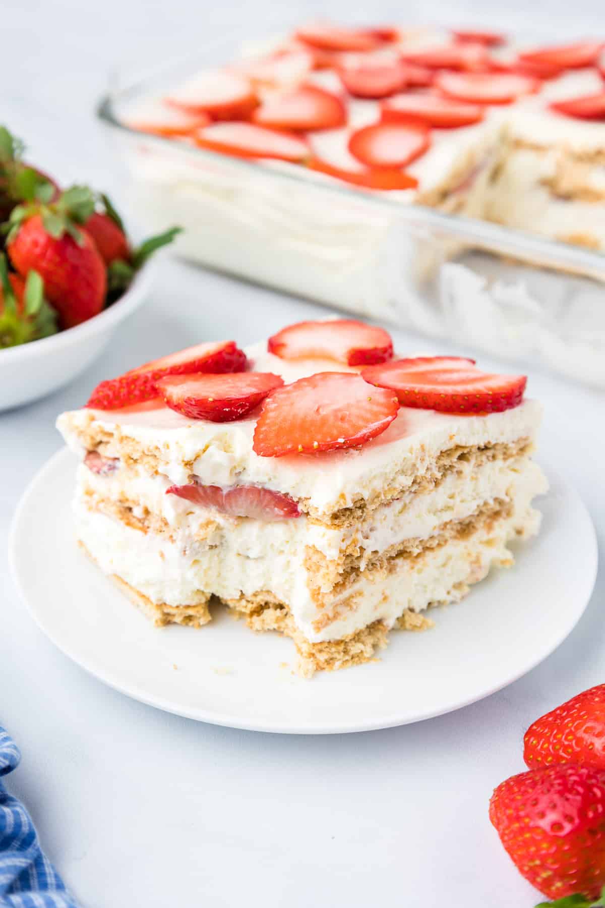 A slice of strawberry cream cheese icebox cake on a plate missing a bite with the rest of the cake and more strawberries in the background on the counter.