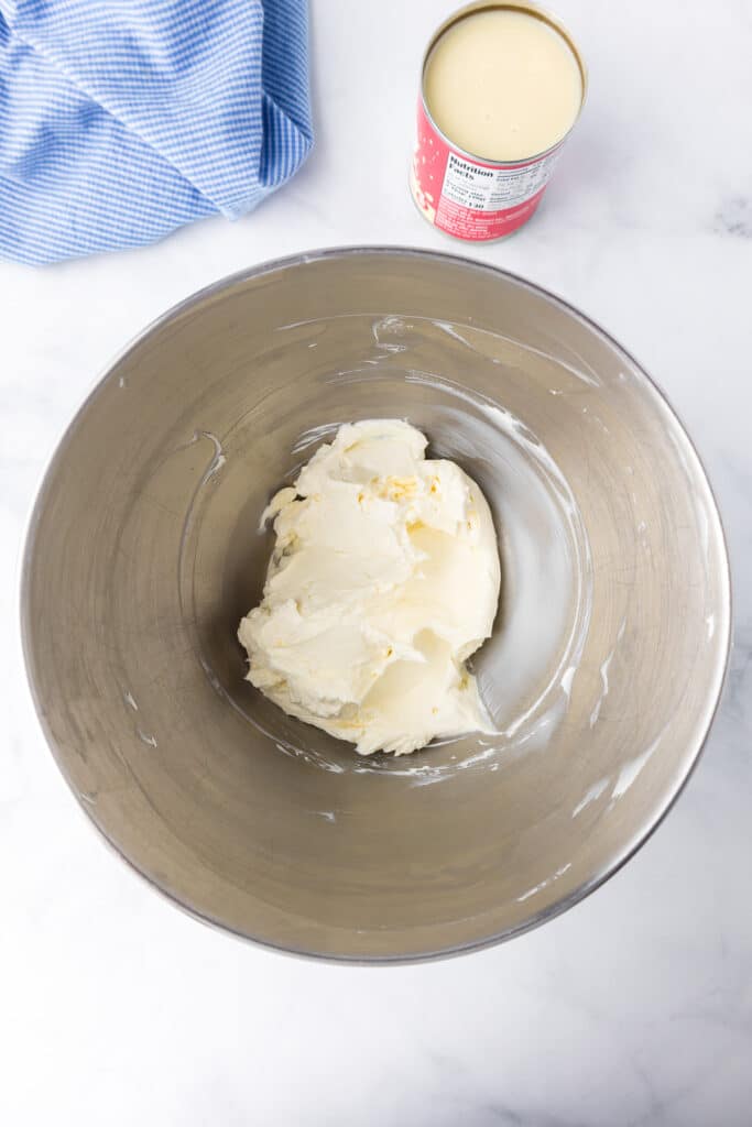 Cream cheese in a large mixing bowl from overhead with an open can of sweetened condensed milk next to the bowl on the counter.