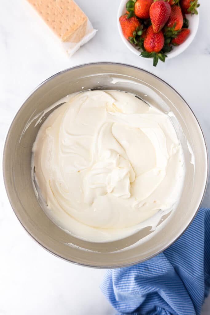 Whipped topping after being folded into the cream m mixture with graham cracker and strawberries on the counter nearby.