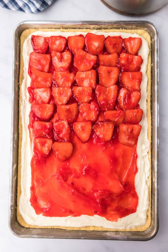Glazed strawberries being layered over strawberry glaze, cream cheese frosting and sugar cookie on a sheet pan from overhead.