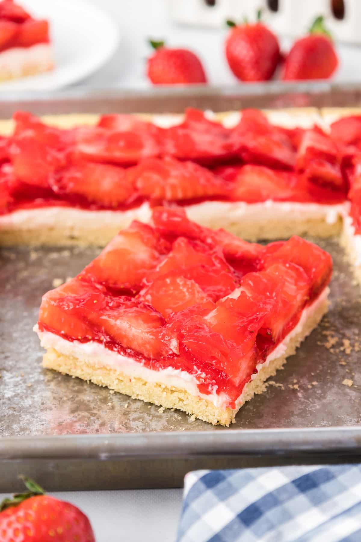 A square slice of strawberry pizza cookie angled on a sheet pan covered in glazed strawberries with more pieces on the pan in the background on a counter.
