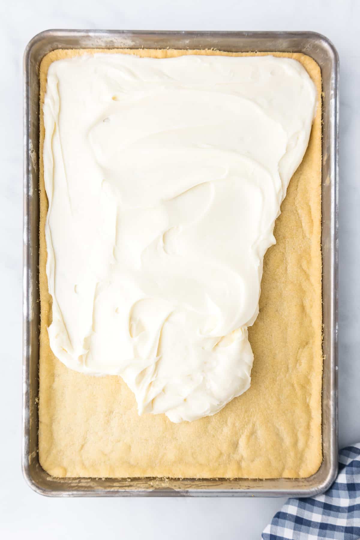 Cream cheese frosting being spread over baked sugar cookie on a sheet pan.