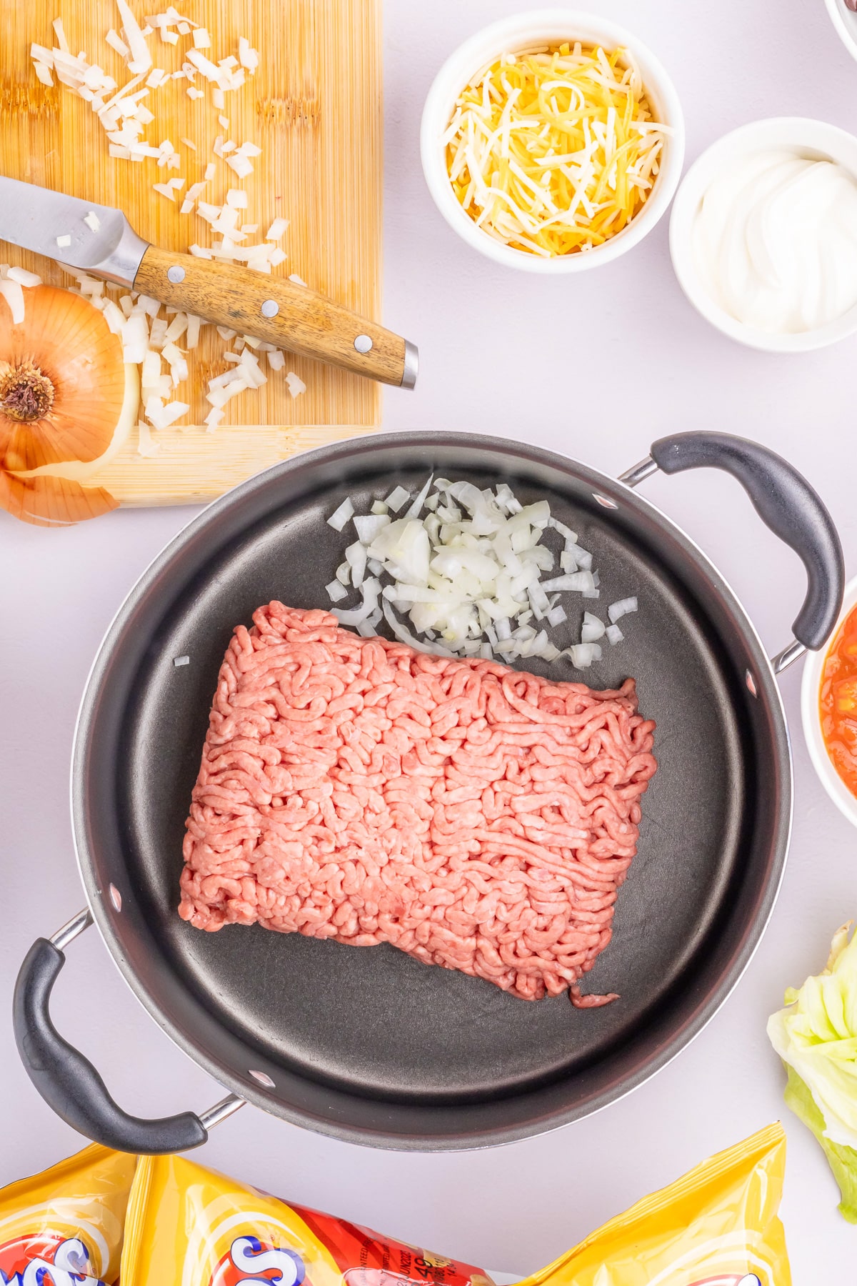 Ground beef and onions in a pan with a cutting board with onions, and bowls of cheese and sour cream nearby on the counter from overhead.
