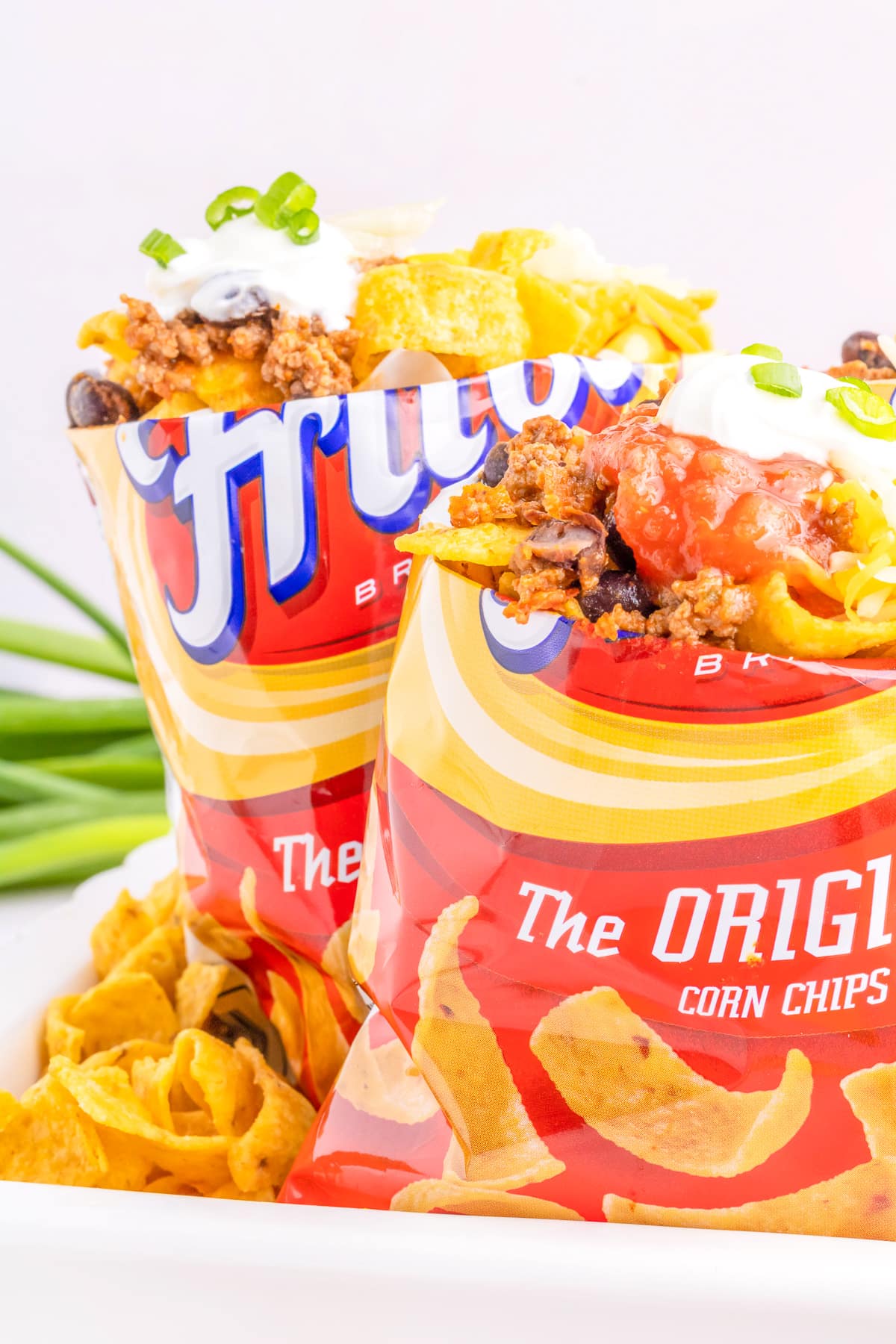 Two bags of Fritos corn chips wit the tops of the bags folded back full of taco meat, salsa and sour cream on top of the chips.