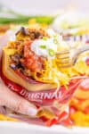 A hand holds a bag of corn chips topped with taco toppings while a fork digs in to the bag to grab a bite.