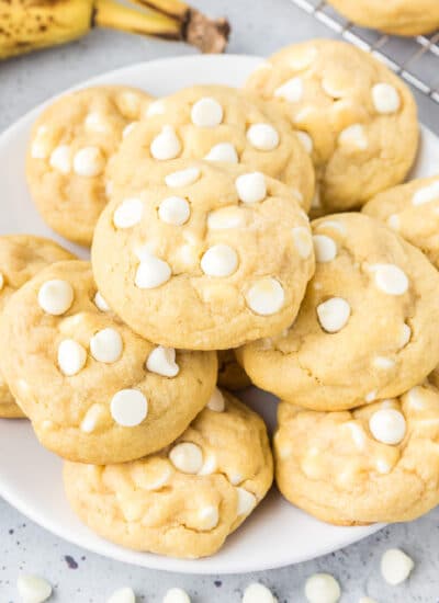 Square view of a plate of banana pudding cookies topped with white chocolate chips on a counter with a banana and a wire cooling rack in the background.